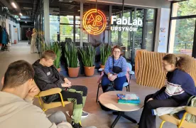 Grupa uczniów siedzi przy stoliku w przestrzeni FabLab Ursynów. W tle widoczne są zielone rośliny w doniczkach oraz neonowe logo FabLab. Uczniowie rozmawiają i odpoczywają w nowoczesnym, jasnym wnętrzu.