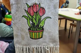 Na zdjęciu widać sylwetke dziecka trzymające uszyt a materiałową torbę. Na środku torby znajduje się kolorowy rysunek przedstawiający czerwone tulipany w doniczce. Nadruk został pokolorowany flamastrami. Torba ma jasny kolor oraz ozdobne frędzle na dole. 