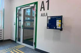 Korytarz szkolny z wejściem do auli A1, oznaczonym dużymi literami. Przed drzwiami znajduje się żółta ścieżka dotykowa prowadząca do wejścia.