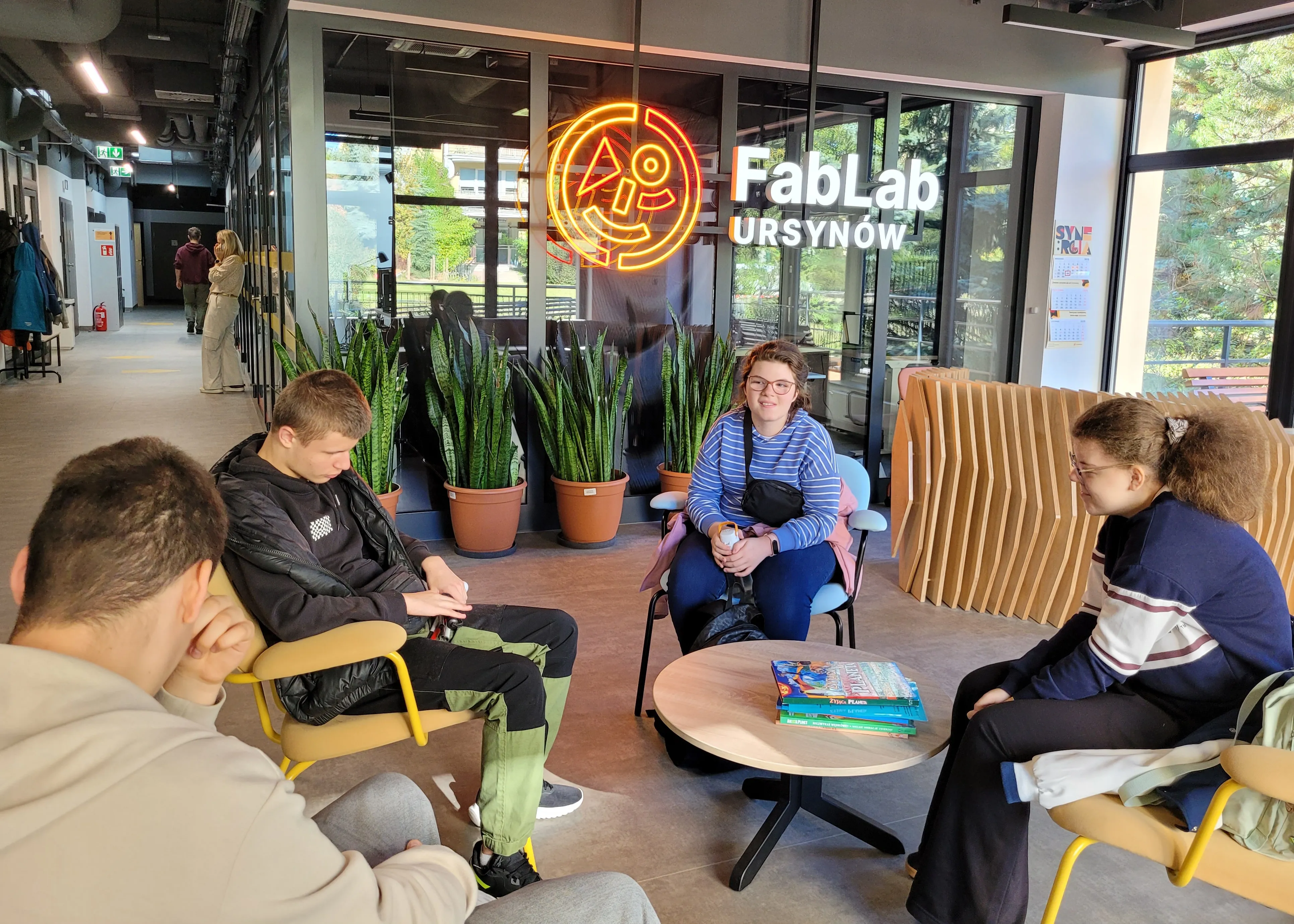 Grupa uczniów siedzi przy stoliku w przestrzeni FabLab Ursynów. W tle widoczne są zielone rośliny w doniczkach oraz neonowe logo FabLab. Uczniowie rozmawiają i odpoczywają w nowoczesnym, jasnym wnętrzu.