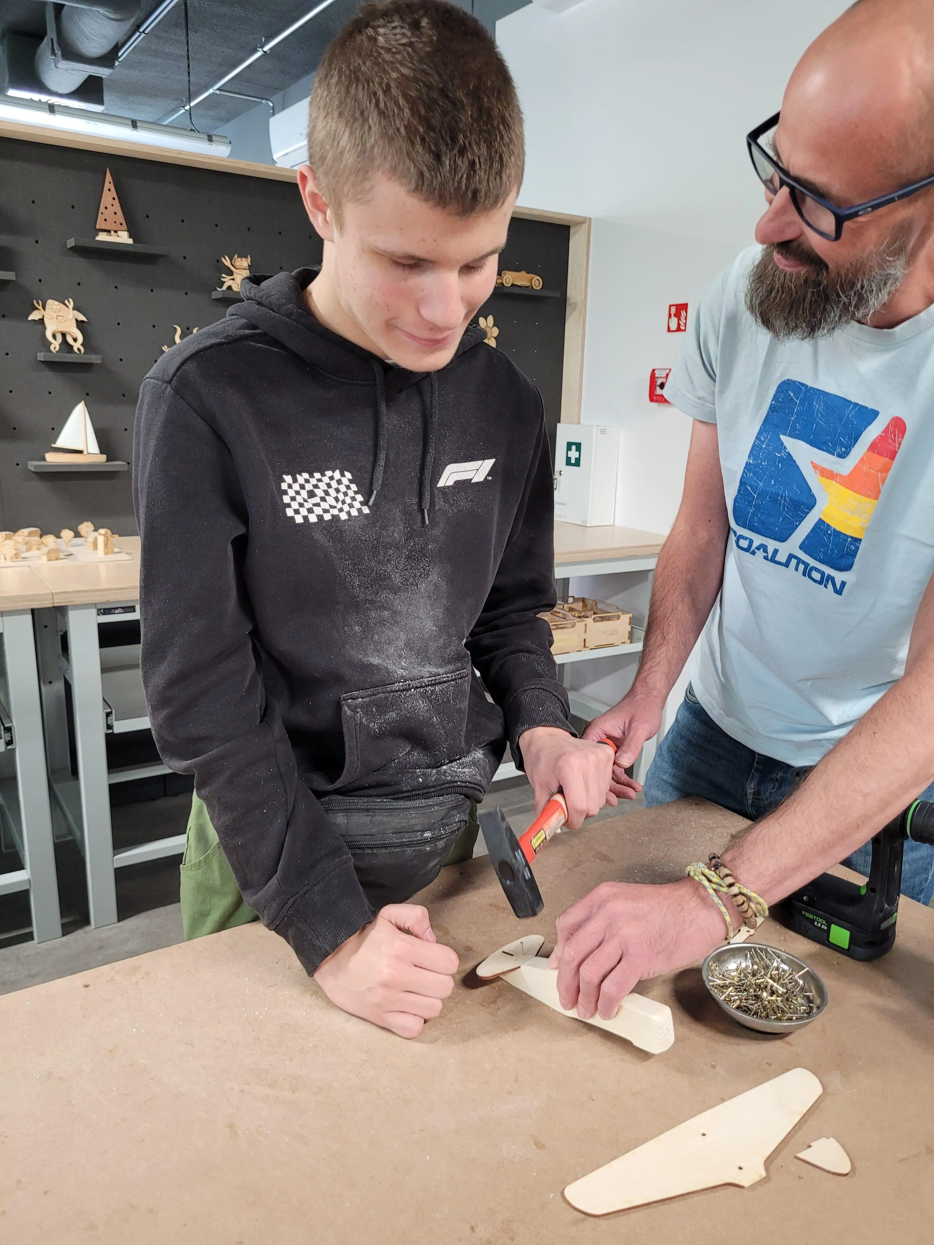 Uczeń w czarnej bluzie wbija gwóźdź w drewniany model, obok instruktor w jasnoniebieskiej koszulce z nadrukiem pomaga przy pracy.