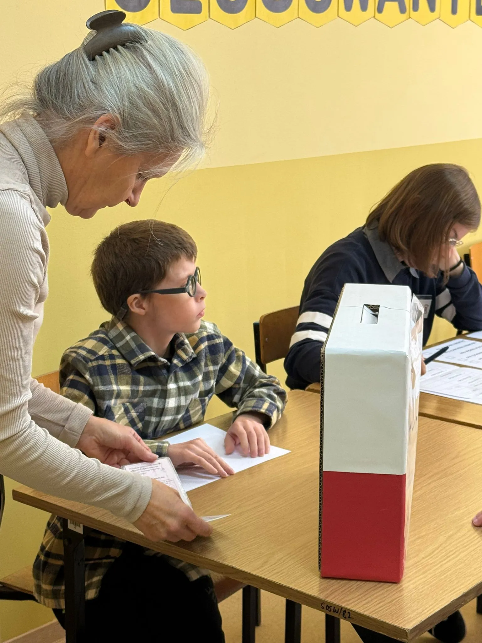 Chłopiec w okularach siedzi przy stole z urną wyborczą, korzystając z karty do głosowania w alfabecie Braille’a. Obok stoi kobieta.