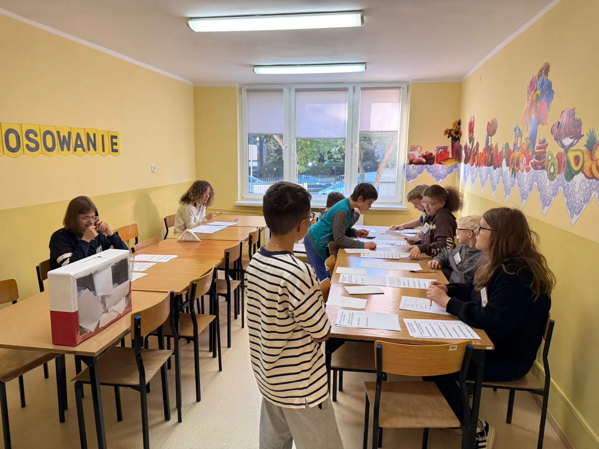 W sali z urną wyborczą uczniowie pełnią rolę komisji, rozdając i sprawdzając karty do głosowania, a inni podchodzą, by oddać głos.
