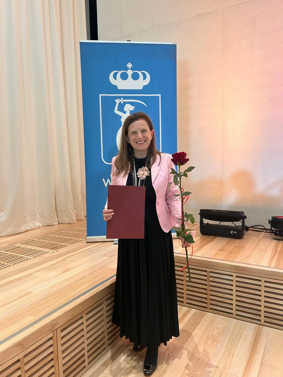 Nauczycielka z dyplomem i czerwoną różą w dłoni po odebraniu Nagrody Dyrektora Biura Edukacji. W tle widoczny jest niebieski baner z herbem Warszawy.