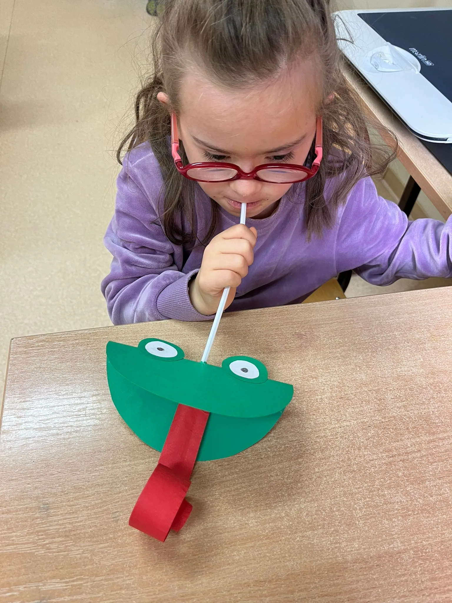 Dziewczynka w okularach siedzi przy biurku i przez słomkę dmucha na papierową żabkę z wystającym czerwonym językiem.