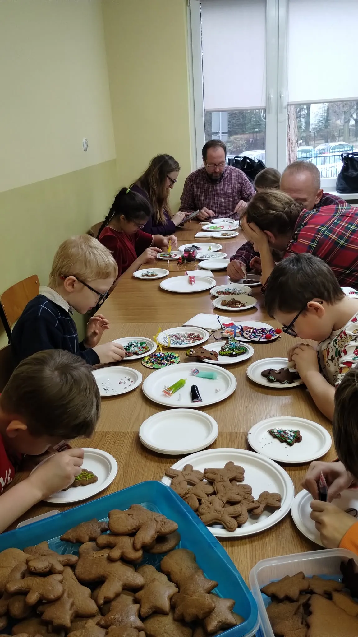 Obraz przedstawia grupę ludzi, w tym dorosłych i dzieci, siedzących wokół prostokątnego drewnianego stołu. Stół jest pokryty różnymi przedmiotami związanymi z dekorowaniem ciasteczek. Na pierwszym planie widoczna jest niebieska taca wypełniona nieozdobionymi piernikami w różnych kształtach, takimi jak gwiazdy czy zwierzęta. Na stole rozłożone są również białe talerze, z których każdy zawiera nieozdobione ciasteczka oraz posypka i lukier. Po obu stronach stołu widać dzieci oraz dorosłych pochylające się do p