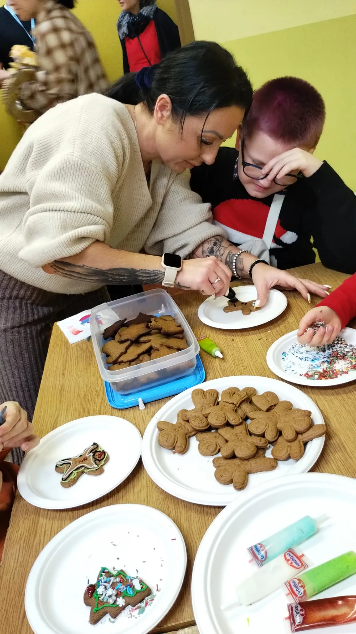 Na zdjęciu widać stół z różnymi talerzami z piernikami i pojemnikami na ciasteczka, a także dwie osoby ściśle ze sobą współpracujące.  Przy ciasteczkach pracują dwie osoby. Kobieta po lewej ma na sobie jasny sweter i inteligentny zegarek na lewym nadgarstku. Pochylają się do przodu pracując nad ozdabianiem pierników. Obok niej siedzi chłopiec w wiśniowych włosach i czarnych okularach. Ma na sobie czarną koszulkę z biało-czerwonym wzorem. Pochyla się do przodu, a jedną rękę ma opartą o stół i podpiera na nie