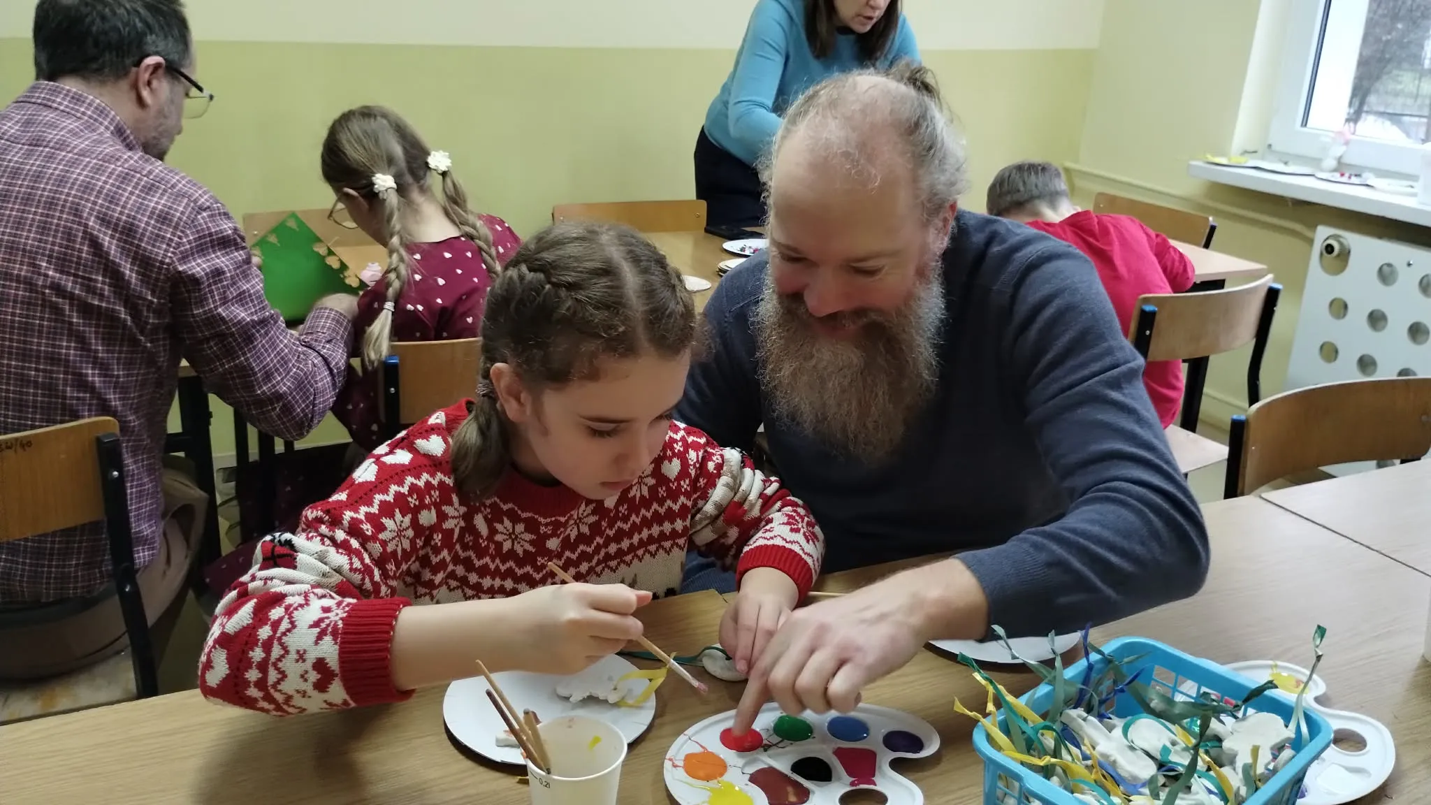 Na zdjęciu dwie osoby siedzą przy stole. Osoba po lewej to młoda dziewczyna ubrana w czerwony sweter z białymi wzorami. Skupia się na malowaniu farami świątecznych ozdób. Przed nią stoi paleta z wieloma kolorami farb, a na stole widoczna jest biała płytka.  Osoba po prawej to dorosły mężczyzna z brodą. Ma na sobie ciemny sweter i ściśle wspiera dziewczynę w jej aktywności. W tle przy stołach siedzą inni ludzie.