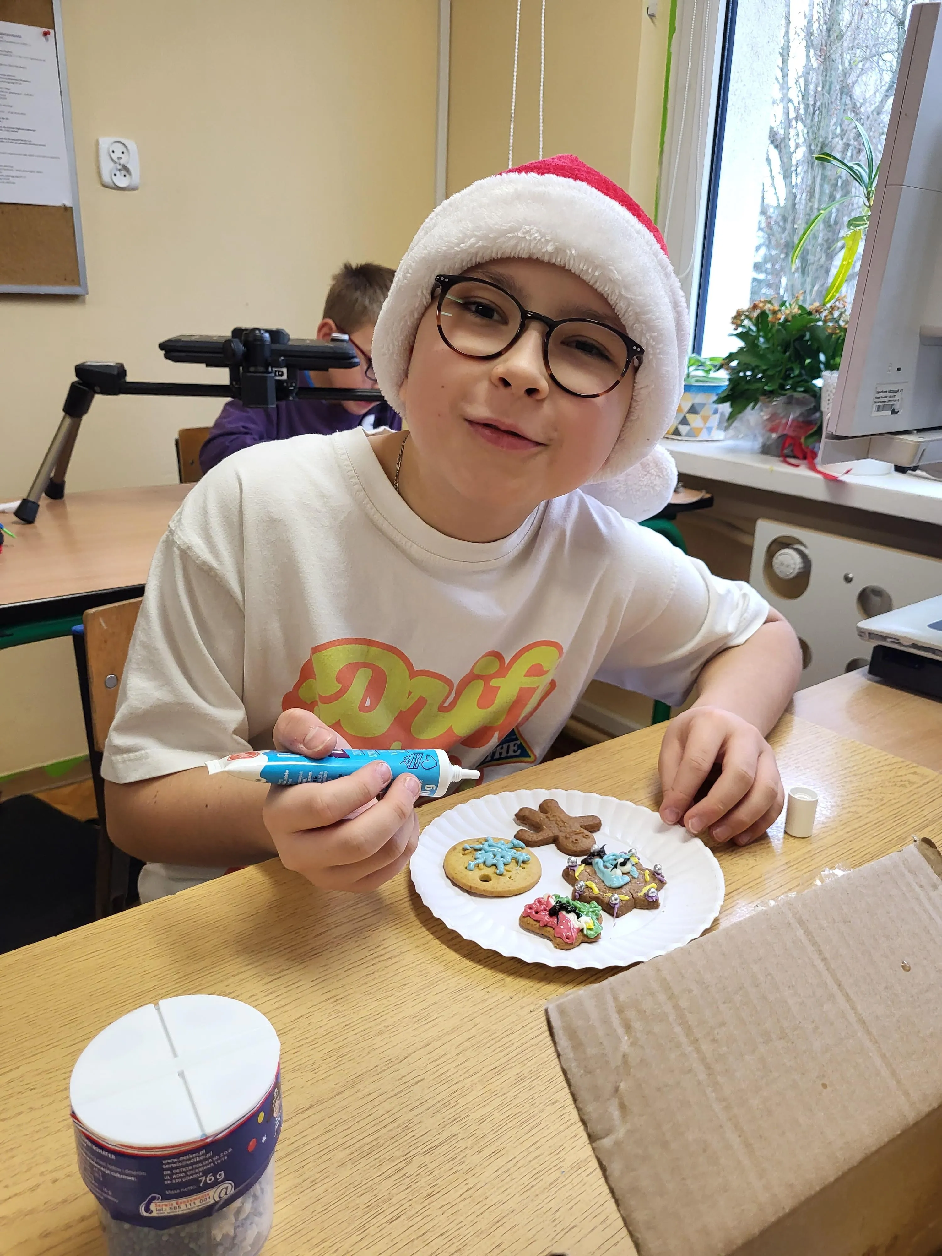 Obraz przedstawia chłopca ubranego w czapkę św. Mikołaja dekorującego świąteczne pierniki kolorowym lukrem