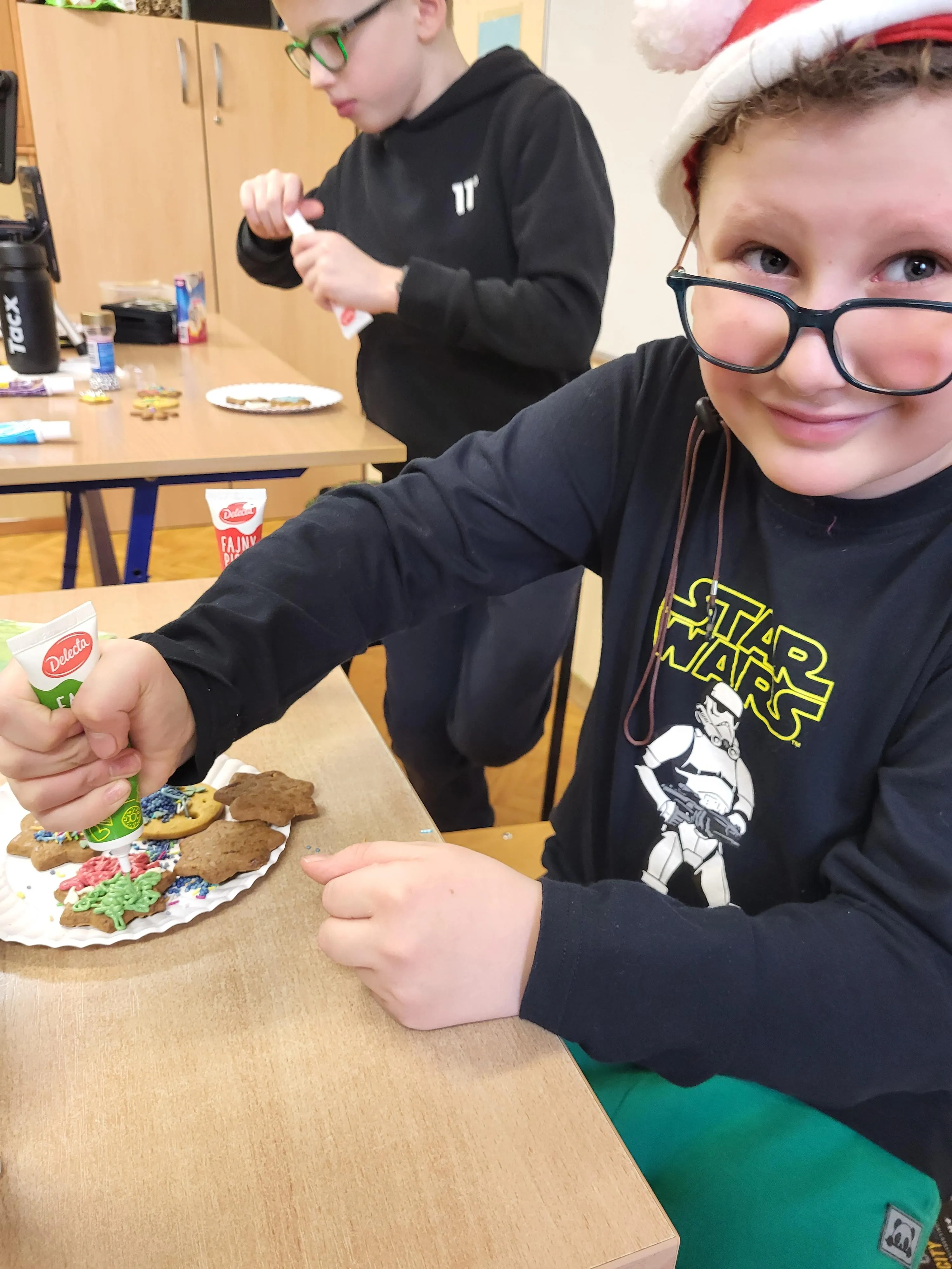 Obraz przedstawia chhłopca ubranego w czapkę św. Mikołaja dekorującego świąteczne pierniki kolorowym lukrem