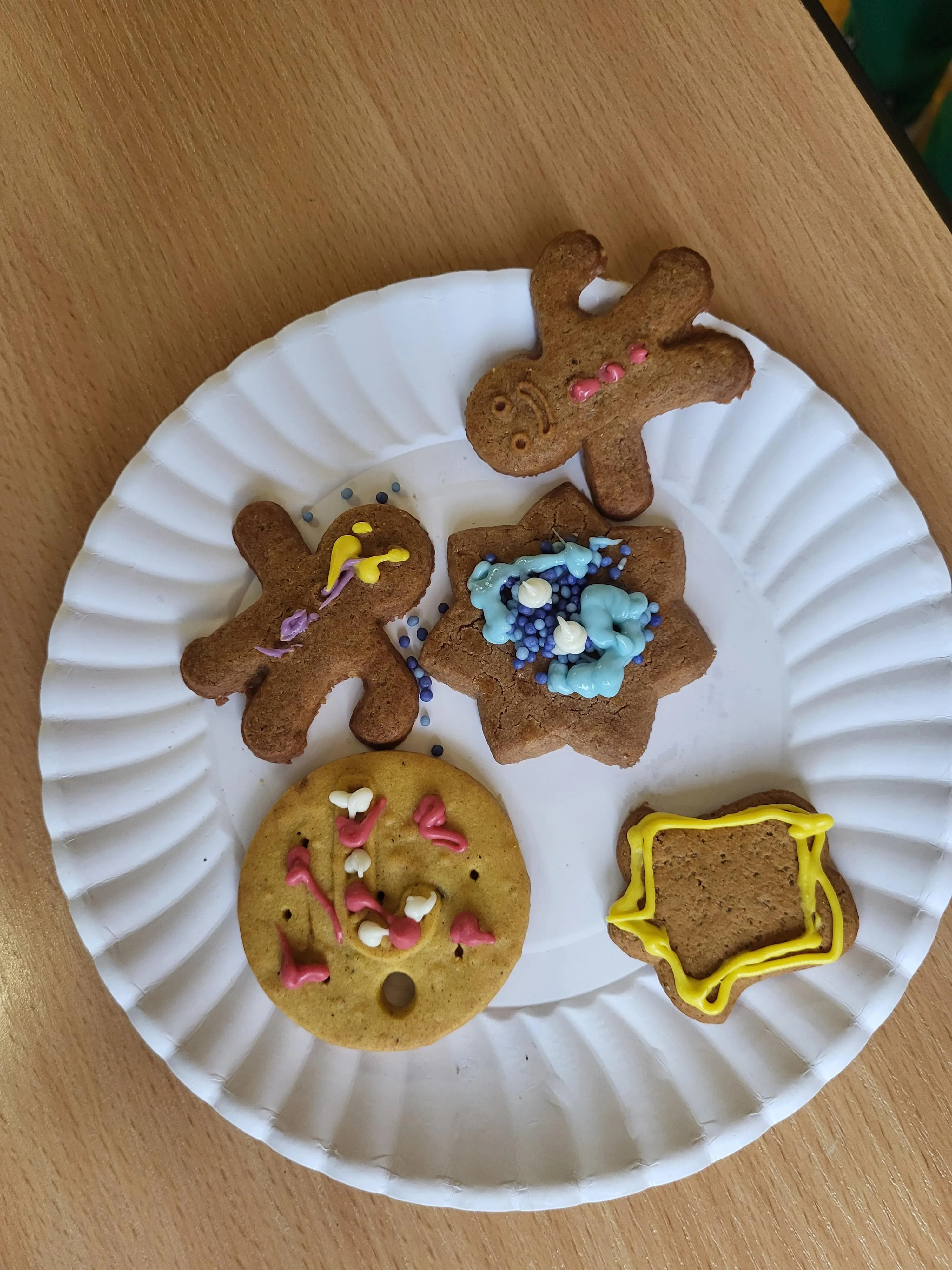 Na obrazie przedstawiono pierniki leżące na białym papierowym talerzu. Ciasteczka są ozdobione kolrowym lukrem oraz jadalnymi ozdobami