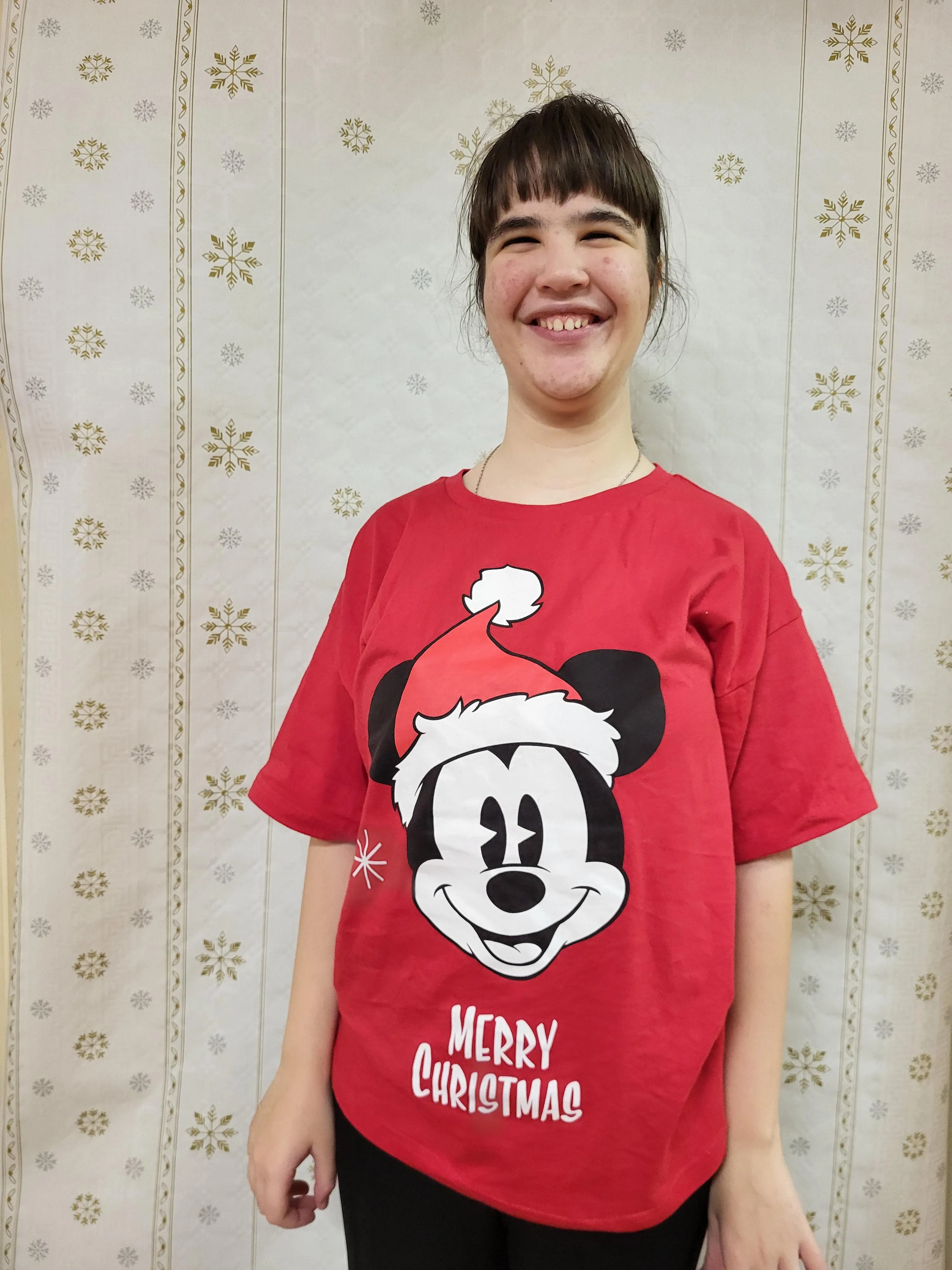 Na zdjęciu widoczna jest uśmiechęnięta dziewczyna w brązowych włosach. Ubrana jest w czerwoną koszulkę na krótki rękaw, na której widnieje duża grafika Myszki Miki w czerwonej mikołajowej czapce i z napisemm Merry Christams umieszczonym pod spodem