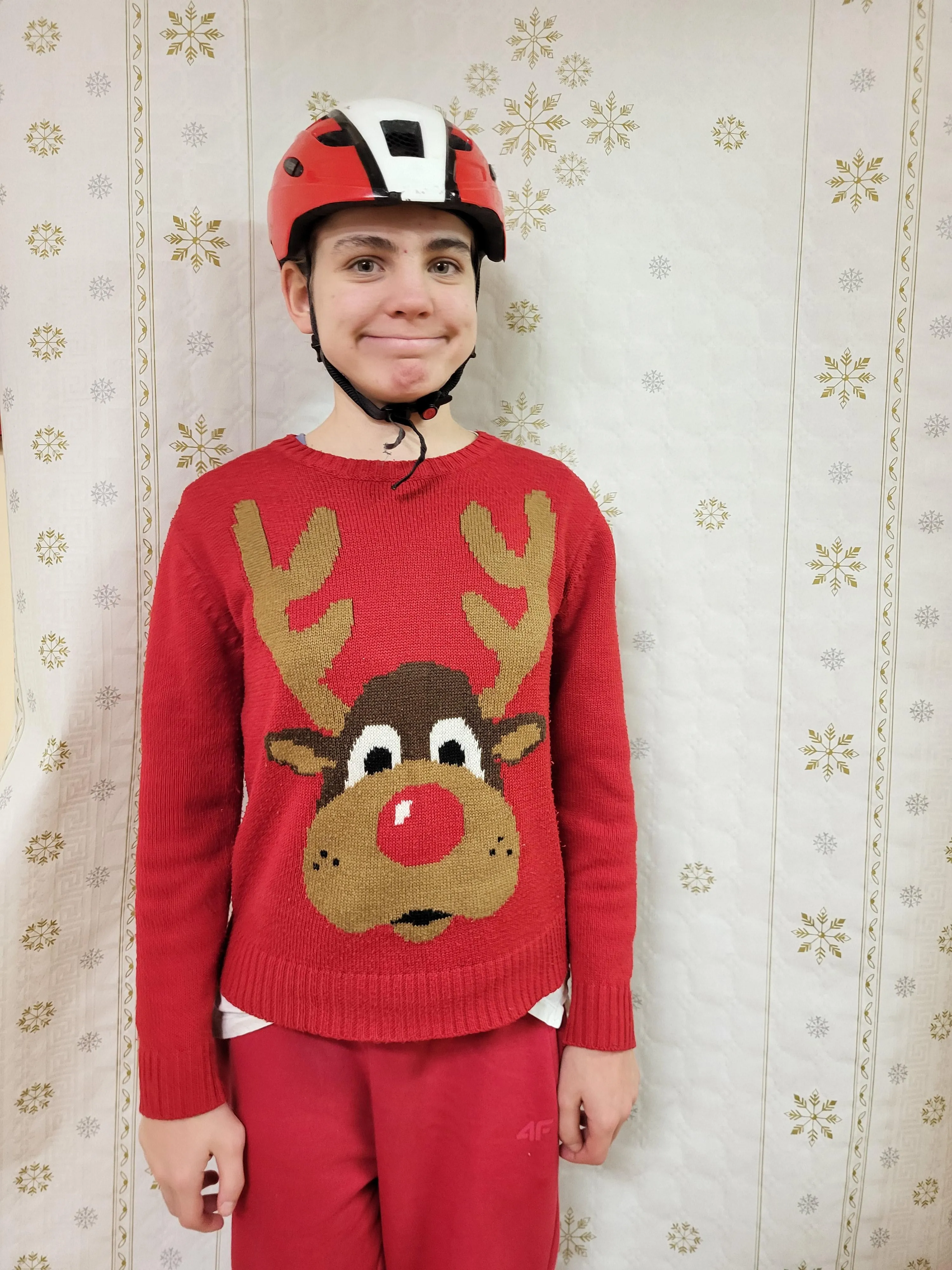 Na zdjęciu widoczna jest uśmiechęnięta dziewczyna w kasku na głowie. Ubrana jest w czerwony sweter z ogromną głową renifera z czerwonym nosem