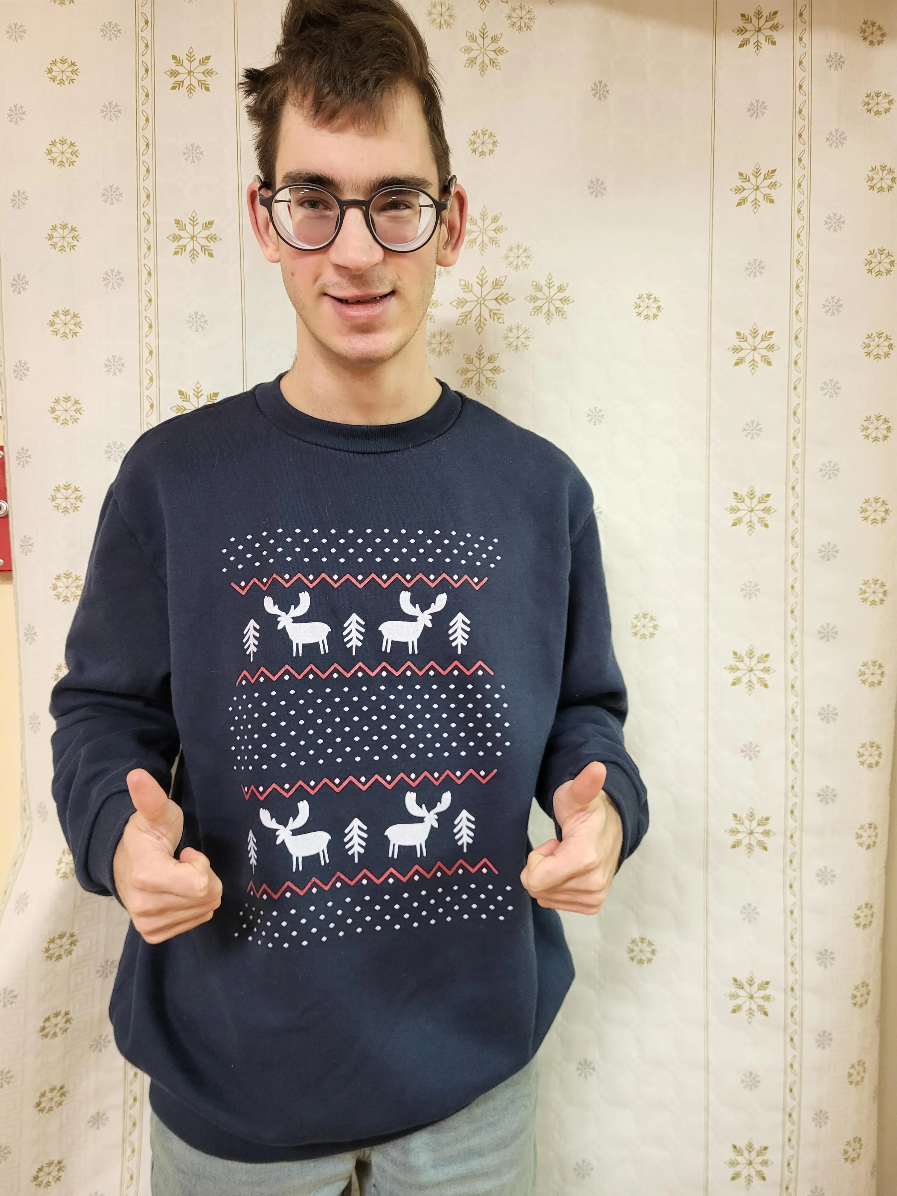 Na zdjęciu widoczny jest uśmiechęnięty chłopiec w brązowych włosach. Ubrany jest w granatowy sweter z białaymi wzorami, reniferami i choinkami