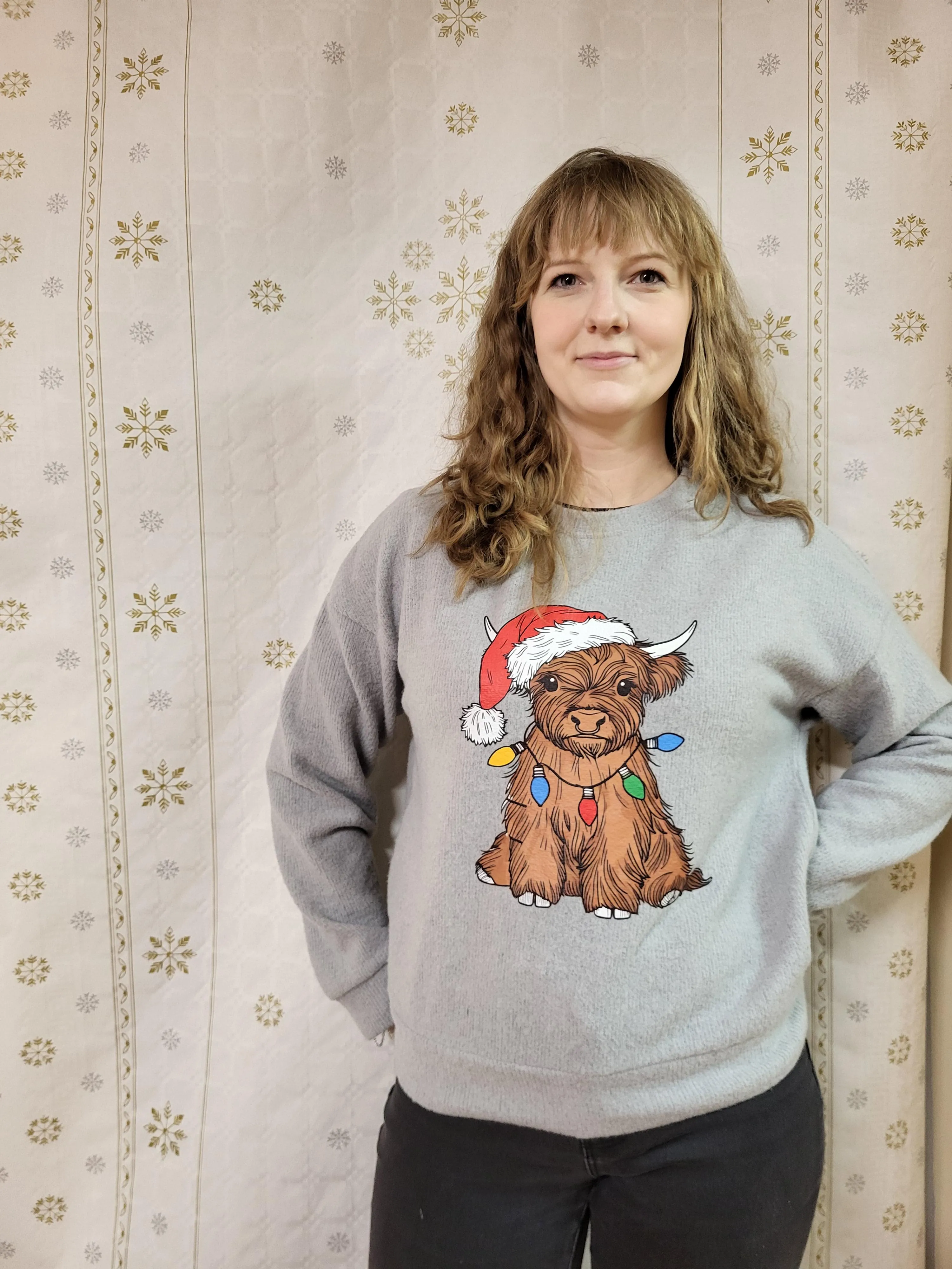 Na zdjęciu widoczna jest uśmiechęnięta kobieta w kręconych blond włosach. Ubrana jest w szary sweter, na któym widnieje siedząca brązowa krowa szkocka ubrana w czepke mikołaja i owinięta w świąteczne lampki dokoła tułowia