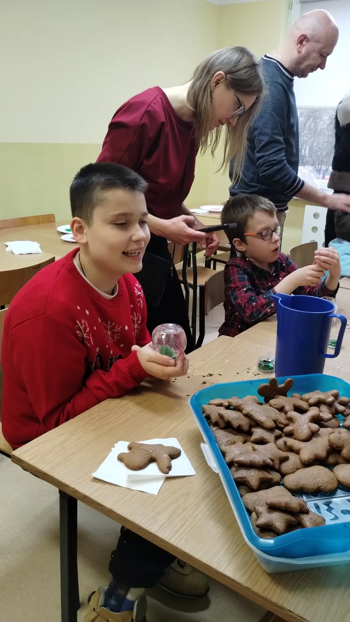 Obraz przedstawia grupę osób zaangażowanych w dekorowanie pierników wokół stołu.  Na pierwszym planie siedzi przy stole chłopiec w czerwonym swetrze z białym wzorem. Trzyma w rękach śnieżną kulę. Przed nim na stole leży mały piernik w kształcie ludzika oraz taca z wieloma piernikami w różnych kształtach. Chłopiec jest uśmiechnięty. Za chłopcem w swetrze siedzi dziecko na krześle, ubrane w koszulę w kratę. Dorosły z długimi włosami, ubrany w czerwoną bluzkę, stoi za dzieckiem i wydaje się mu pomagać. 