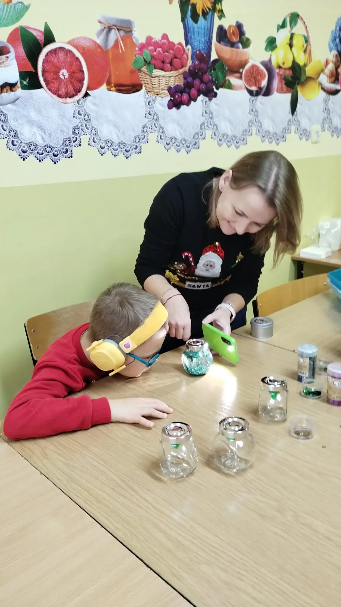 Dziecko w czerwonym swetrze i słuchawkach wyciszających z żółtym paskiem siedzi przy drewnianym stole. Dziecko pochyla się do przodu, wchodząc w interakcję z małymi śnieżnymi kulami leżącymi na stole przed nim. Obok dziecka stoi kobieta, której twarz jest zasłonięta, pomagając mu. Ma na sobie sweter z uroczystym motywem Świętego Mikołaja. 