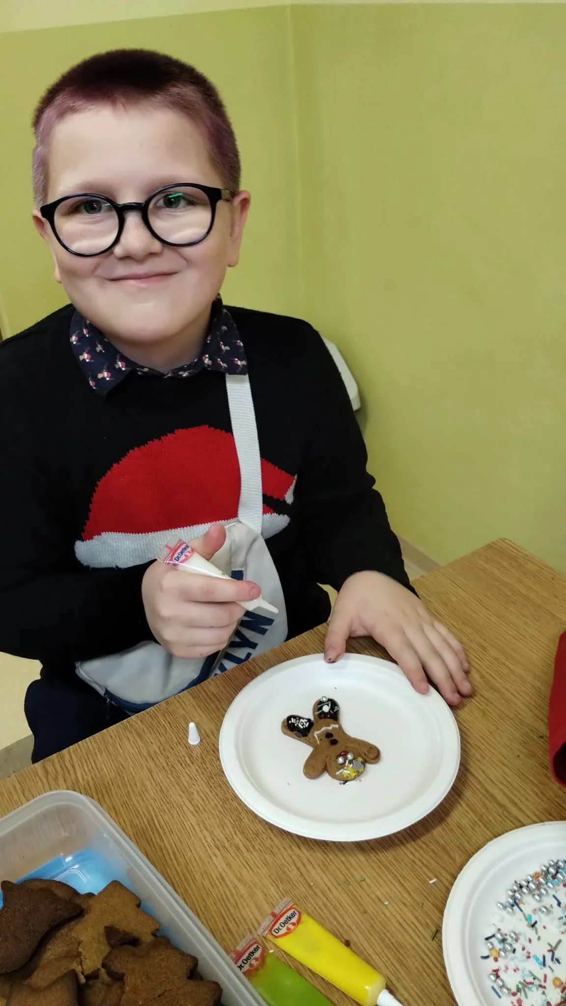 Zdjęcia przedstawia chłopca z wiśniowymi krótkimi włosami stojącego przy stole, ubranego w sweter ze Świętym Mikołajem. Chłopiec trzyma w prawej ręce małą tubkę z lukrem. Na talerzu znajduje się ozdobne piernikowe ciasteczko w kształcie ludzika, który jest już częściowo ozdobiony lukrem i kolorową posypką. Po lewej stronie talerza znajduje się taca z dodatkowymi, nieozdobionymi piernikami. 