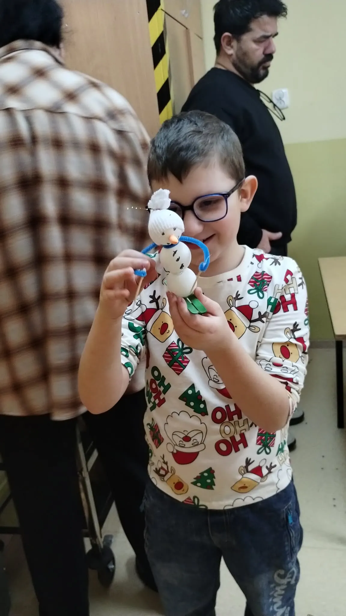 Na zdjęciu widać dziecko trzymające w ręku małego bałwana, jadącego na nartach. Dziecko ma na sobie jeansy oraz  białą koszulkę z krótkim rękawem i kolorowymi motywami świątecznymi, w tym choinkami, reniferami. W tle stoi osoba w koszuli w kratę, częściowo widoczna, oraz inna osoba w ciemnych ubraniach stojąca dalej z tyłu. 