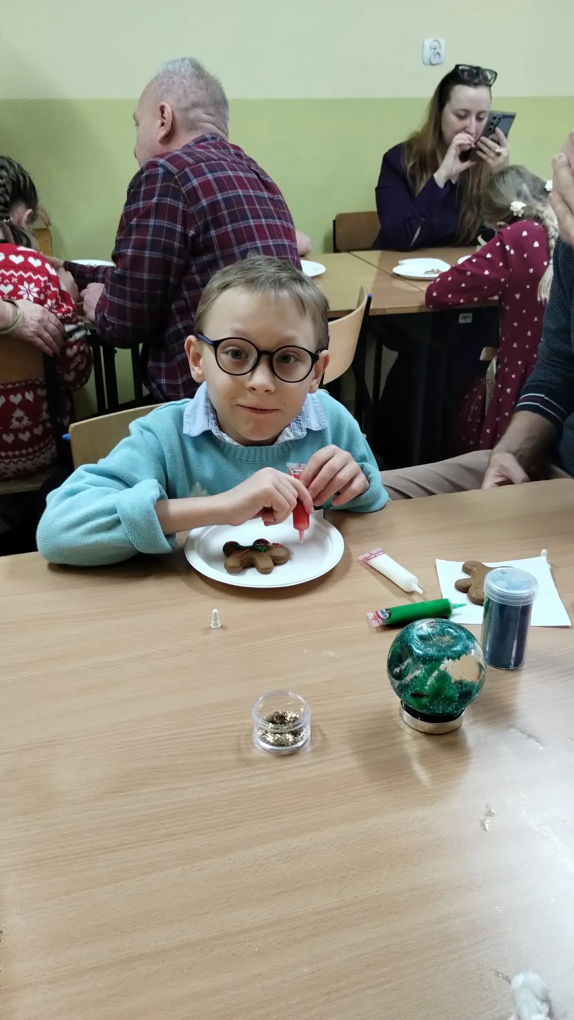 Na zdjęciu widać pomieszczenie z kilkoma osobami siedzącymi przy stołach. Na pierwszym planie widać chłopca w jasnoniebieskiej koszuli, siedzące przy stole. Dziecko pracuje nad talerzem, na którym znajduje się piernik w kształcie ludzika. Na stole przed dzieckiem leży śnieżna kula, pisaki z lukrem oraz kolorowa posypka. W tle stoją dwa stoliki z siedzącymi przy nich ludźmi, siedzącymi tyłem i przodem do kamery. 