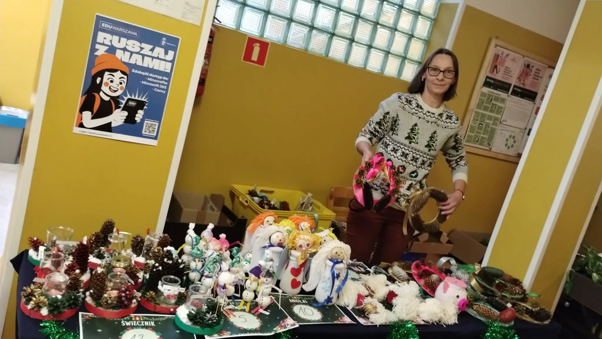 Obraz przedstawia świąteczny kiermasz. Na zajęciu widoczne jest pomieszczenie ze stołem, pokrytym różnymi ozdobnymi przedmiotami, takimi jak małe figurki, ozdoby i kompozycje z szyszek. Przedmioty te są ułożone w grupy, niektóre umieszczone w przezroczystych pojemnikach lub na małych stojakach. Stół jest ustawiony przy żółtej ścianie. Po prawej stronie stołu stoi kobieta, trzymając w rękach dwa wieńce świąteczne. Kobieta ma na sobie okulary, biały sweter z czarnymi wzorami drzew i ciemne spodnie. 