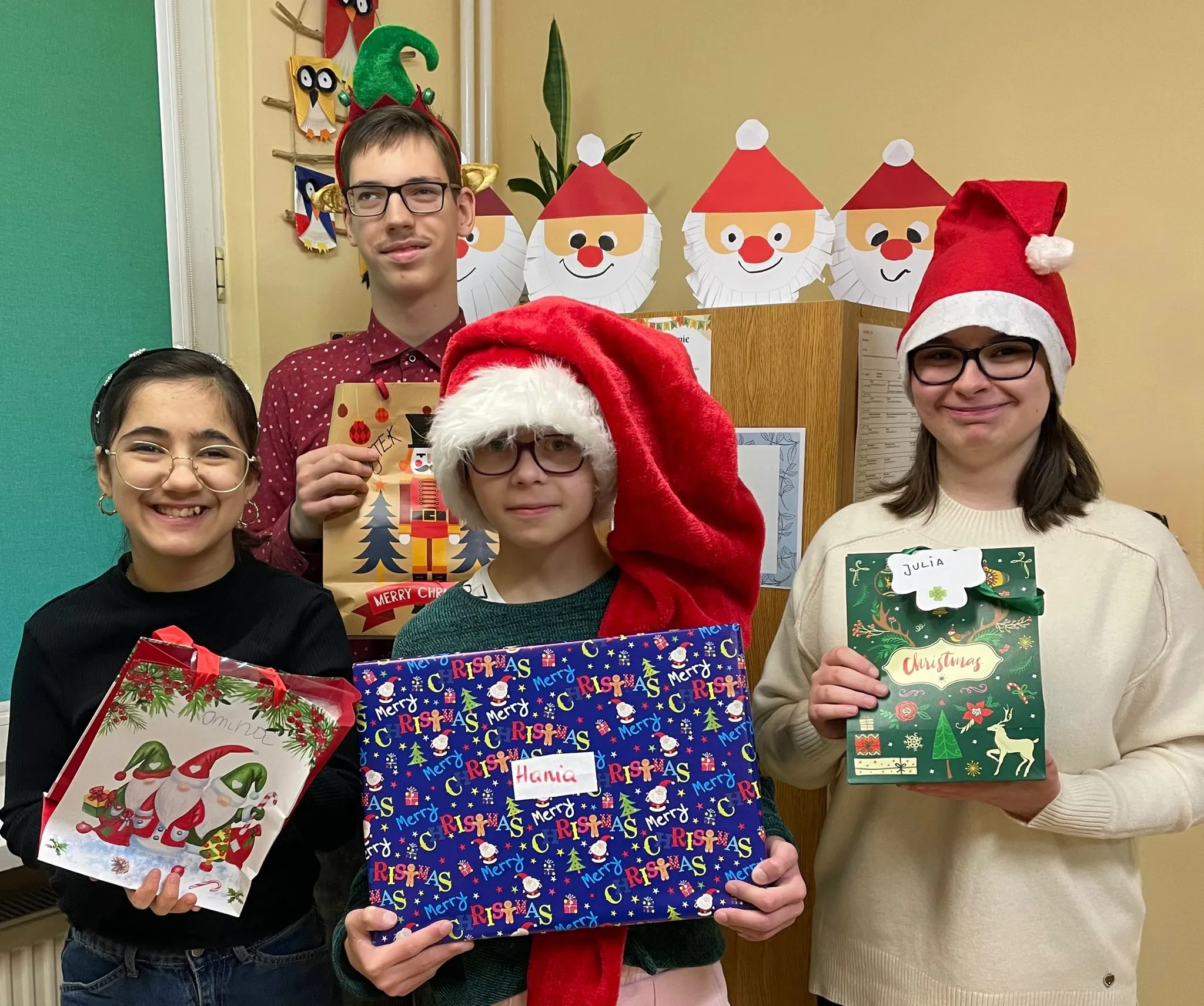 Obraz przedstawia 4 dzieci ustawionych przed ścianą ozdobioną papierowymi twarzami św. Mikołaja. Wszystkie osoby na obrazie uśmiechają się oraz trzymają w rękach prezenty