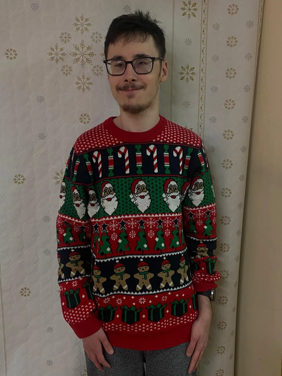 Na zdjęciu widoczny jest uśmiechęnięty chłopiec w brązowych włosach. Ubrany jest w czerwony sweter na którym widnieją poziome pasy ze świętecznymi motywami: cukrowymi laskami, mikołajami, choinkami, piernikami i prezentami