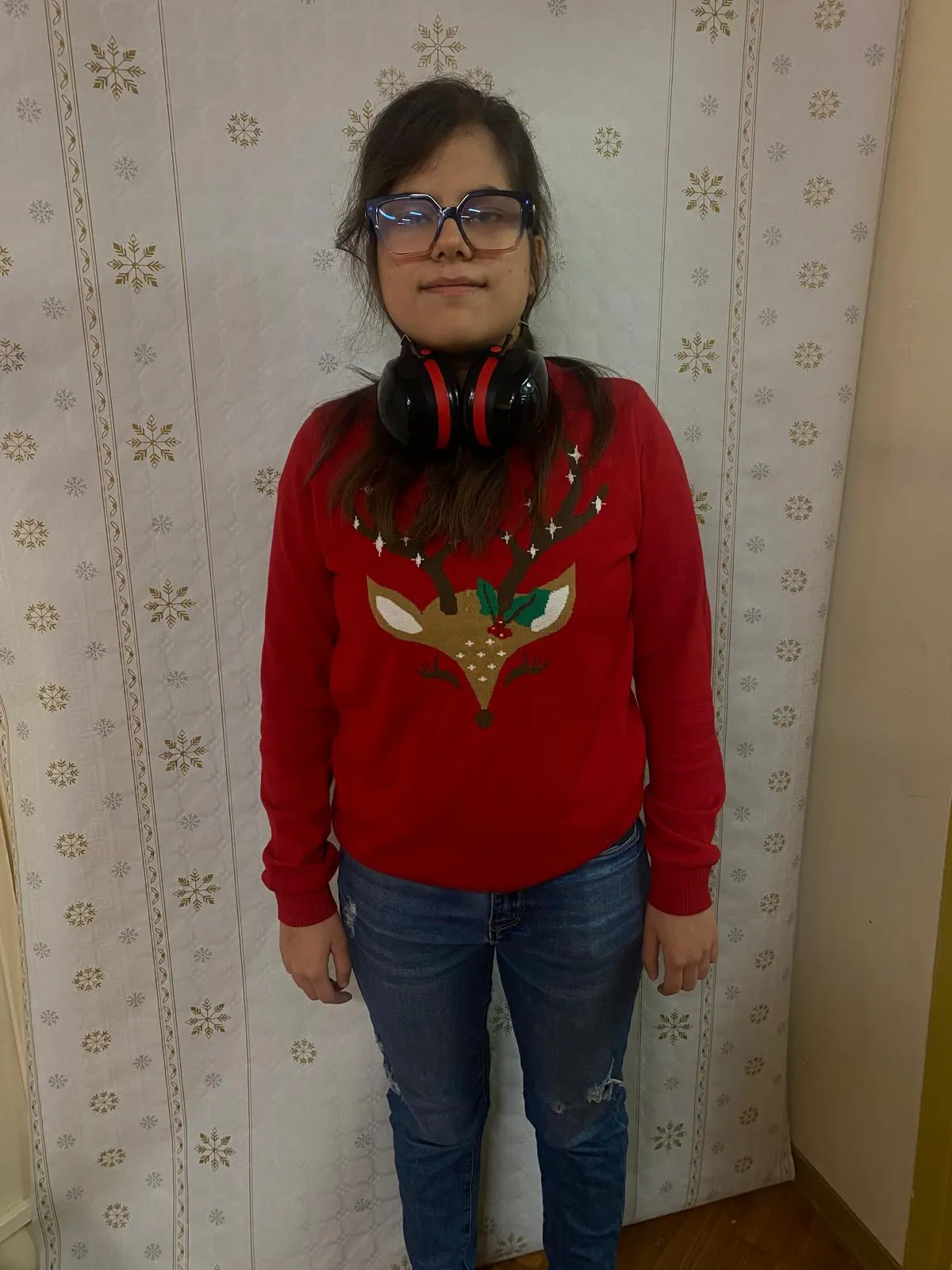 Na zdjęciu widoczna jest uśmiechęnięta dziewczyna w brązowych włosach. Ubrana jest w czerwony sweter z ogromną głową renifera