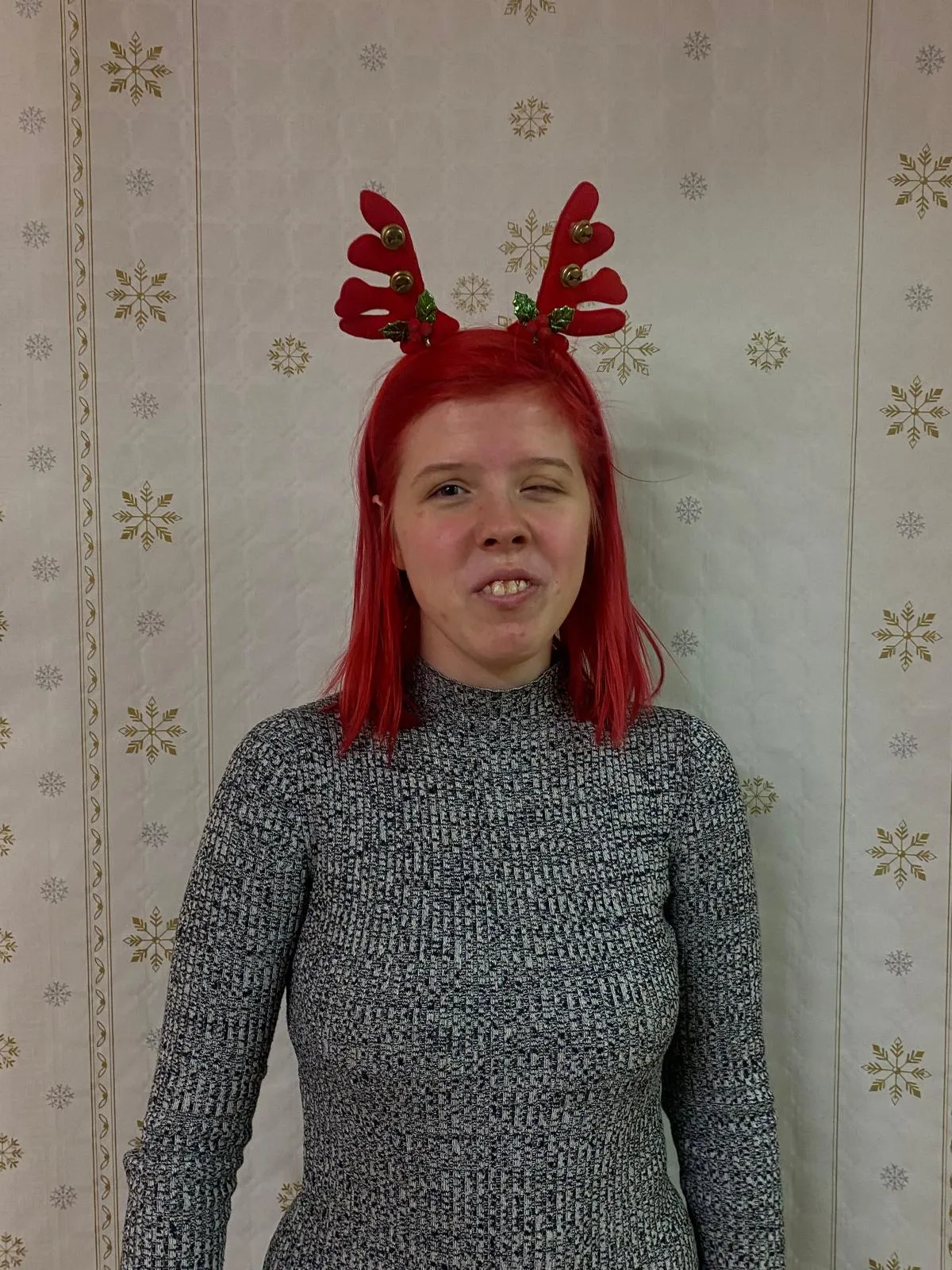 Na zdjęciu widoczna jest uśmiechęnięta dziewczyna w brązowych włosach. Ubrana jest w szarą koszulkę, a na jej głowie widać czerwoną opaskę z rogami renifera