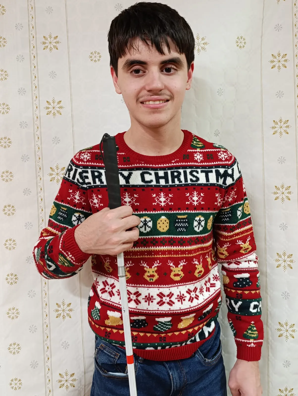 Na zdjęciu widoczny jest uśmiechęnięty chłopiec w brązowych włosach trzymający w ręku białą laskę. Ubrany jest w czerwony sweter na którym widnieją poziome pasy ze świętecznymi motywami: reniferami, bombkami, skarpetami, choinkami. Na swetrze widnieje napis Merry Christmas