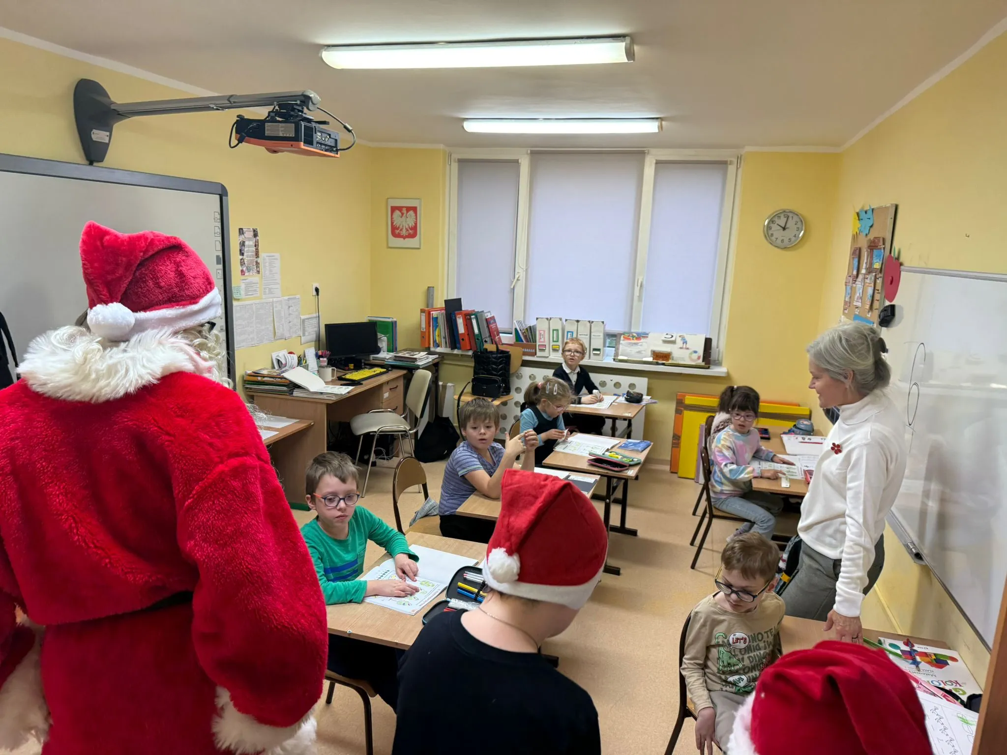Obraz przedstawia klasę z kilkoma dziećmi siedzącymi przy ławkach oraz osobą ubraną w strój Św. Mikołaja