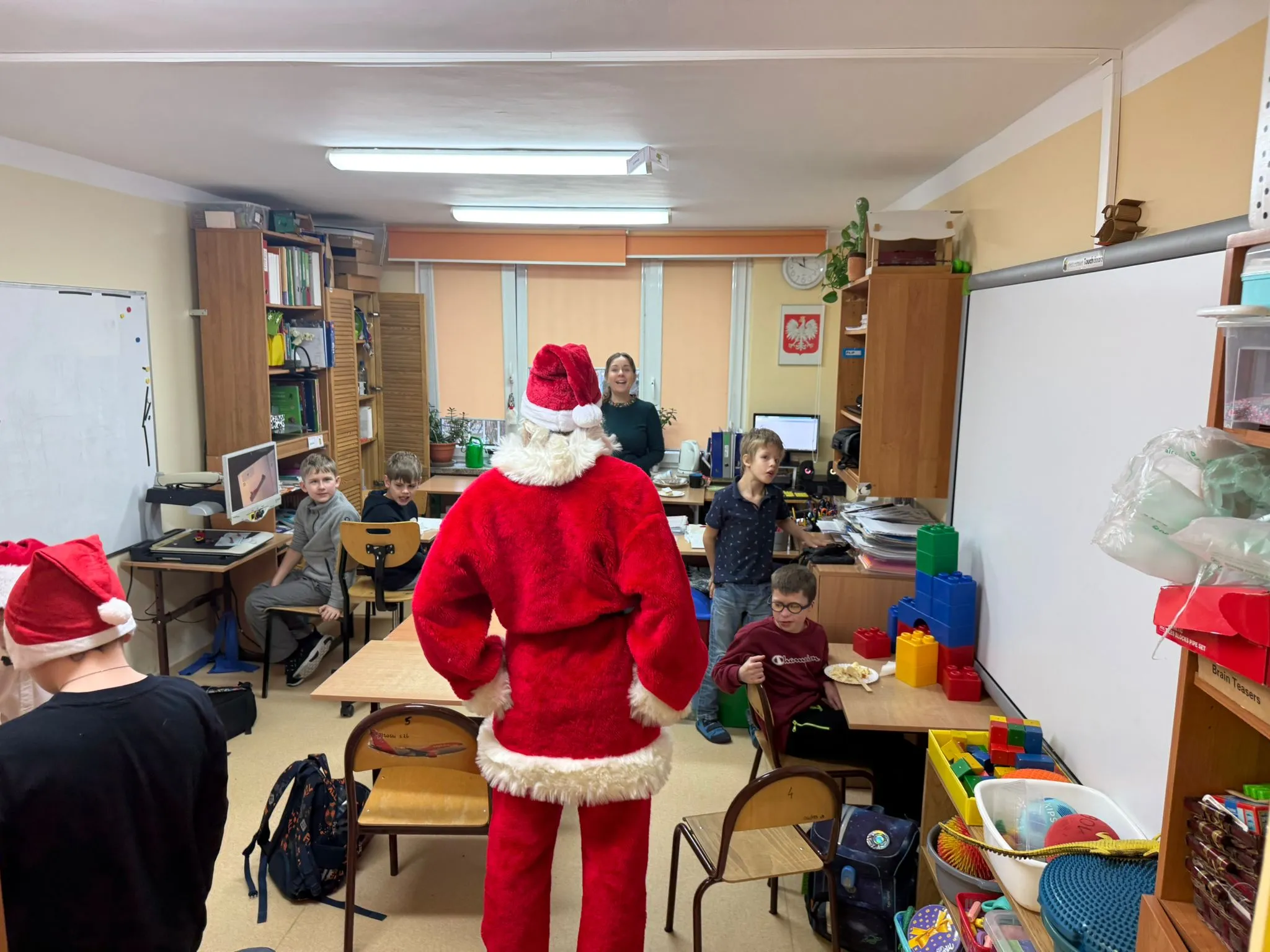 Obraz przedstawia klasę z kilkoma dziećmi siedzącymi przy ławkach oraz osobą ubraną w strój Św. Mikołaja stojącą pod tablicą szkolną