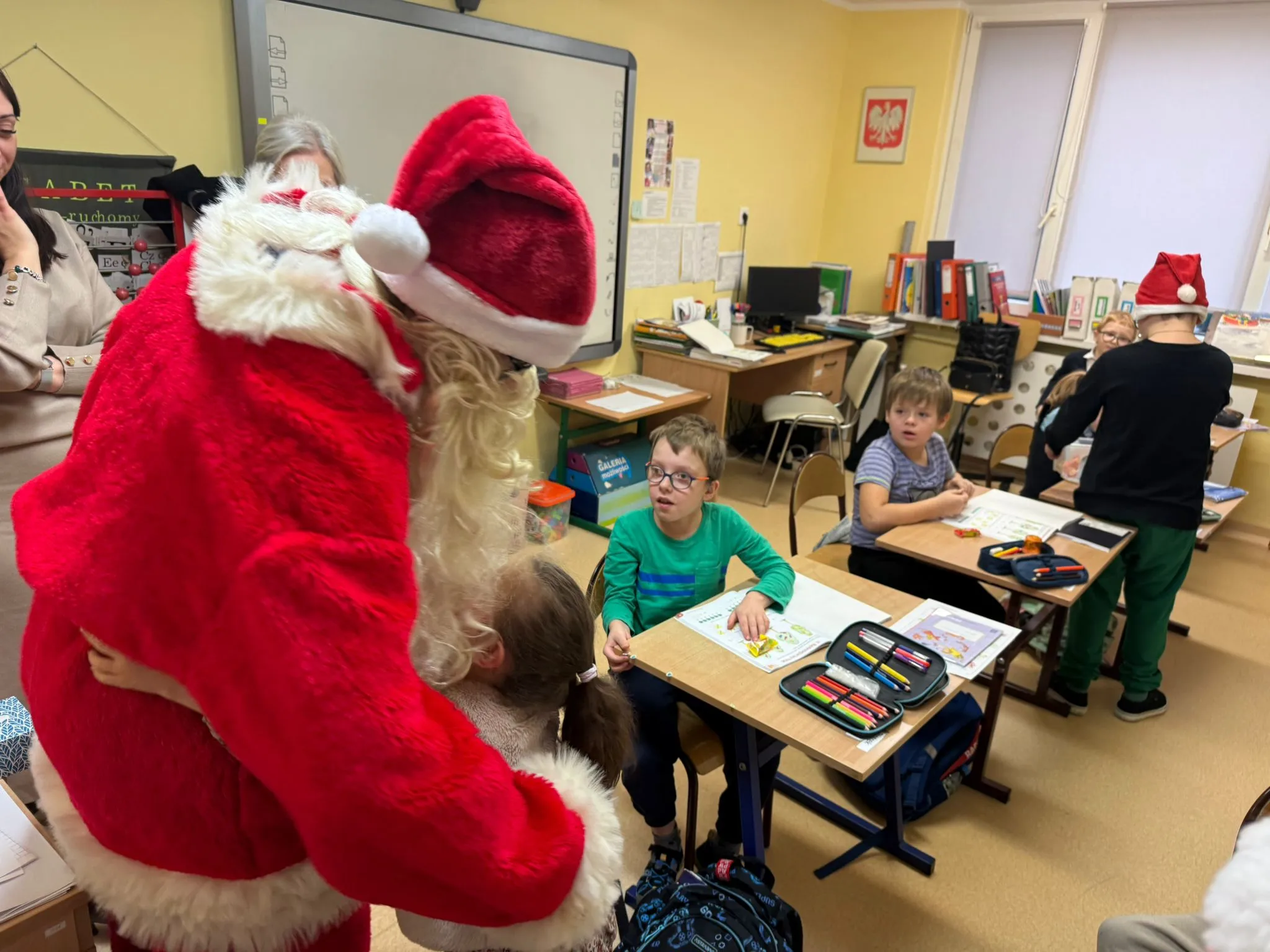 Obraz przedstawia klasę z kilkoma dziećmi siedzącymi przy łąwkach oraz osoą ubraną w strój Św. Mikołaja, który przytula jedno z dzieci. 