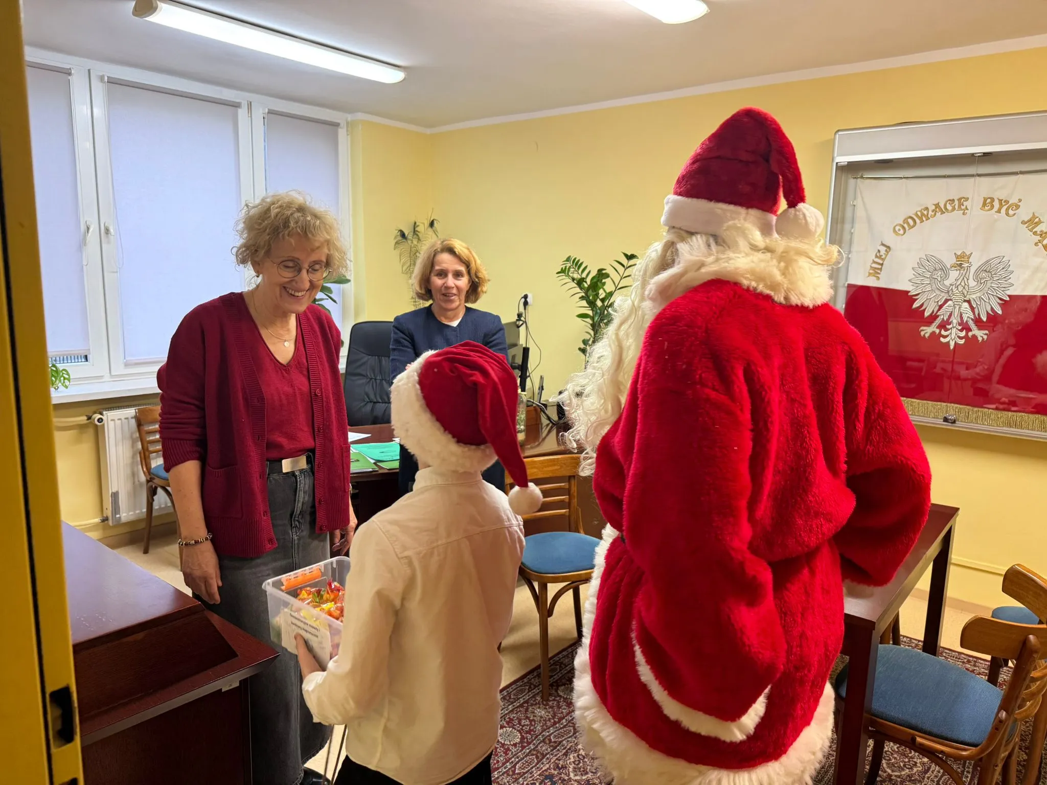 Obraz przedstawia osobę przebraną za św. Mikołaja oraz osobe przebraną za świątecznego Elfa, wręczajce słodkości dwóm kobietą - dyrektorce Ośrodka oraz kierowniczce Internatu 