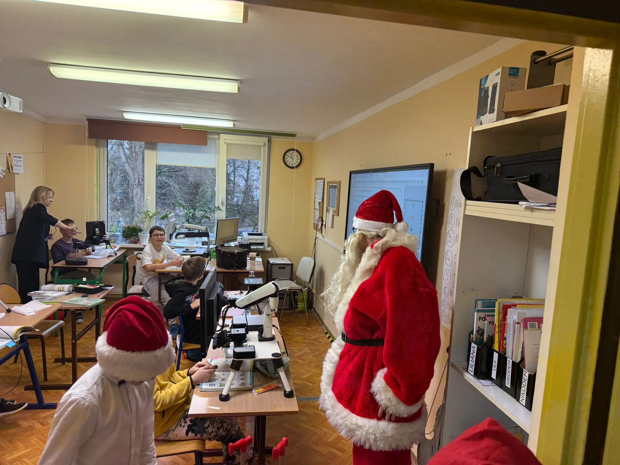 Obraz przedstawia klasę z kilkoma dziećmi siedzącymi przy ławkach oraz osobą ubraną w strój Św. Mikołaja