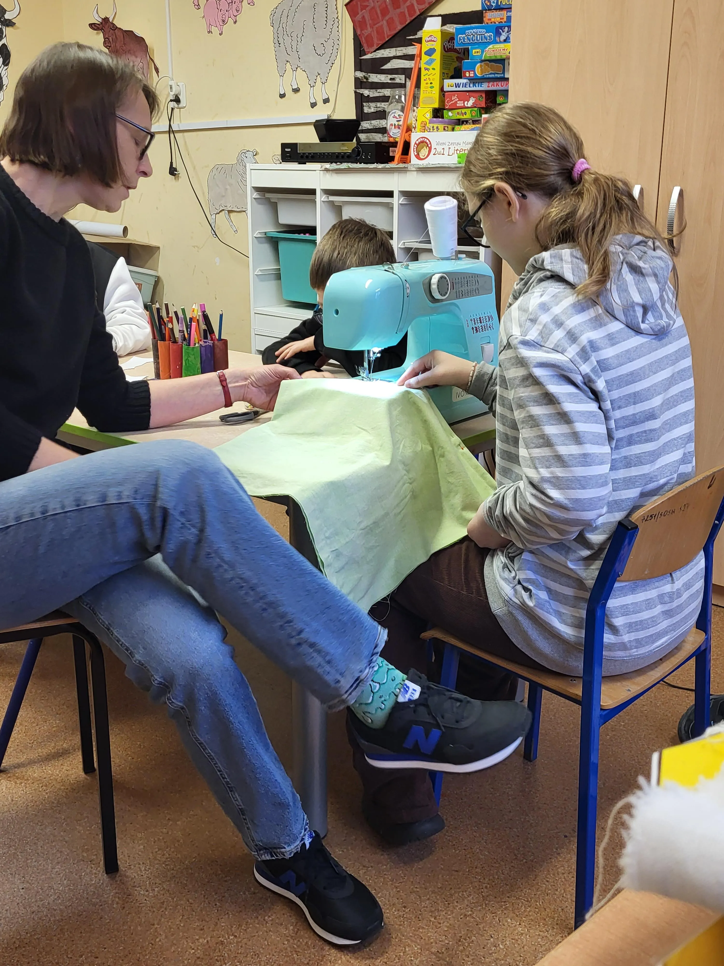 Na zdjęciu widać nauczycielkę i uczennicę pracujące przy maszynie do szycia. Razem szyją materiał, z którego powstaje torba. W tle widać przybory plastyczne oraz szkolne szafki.