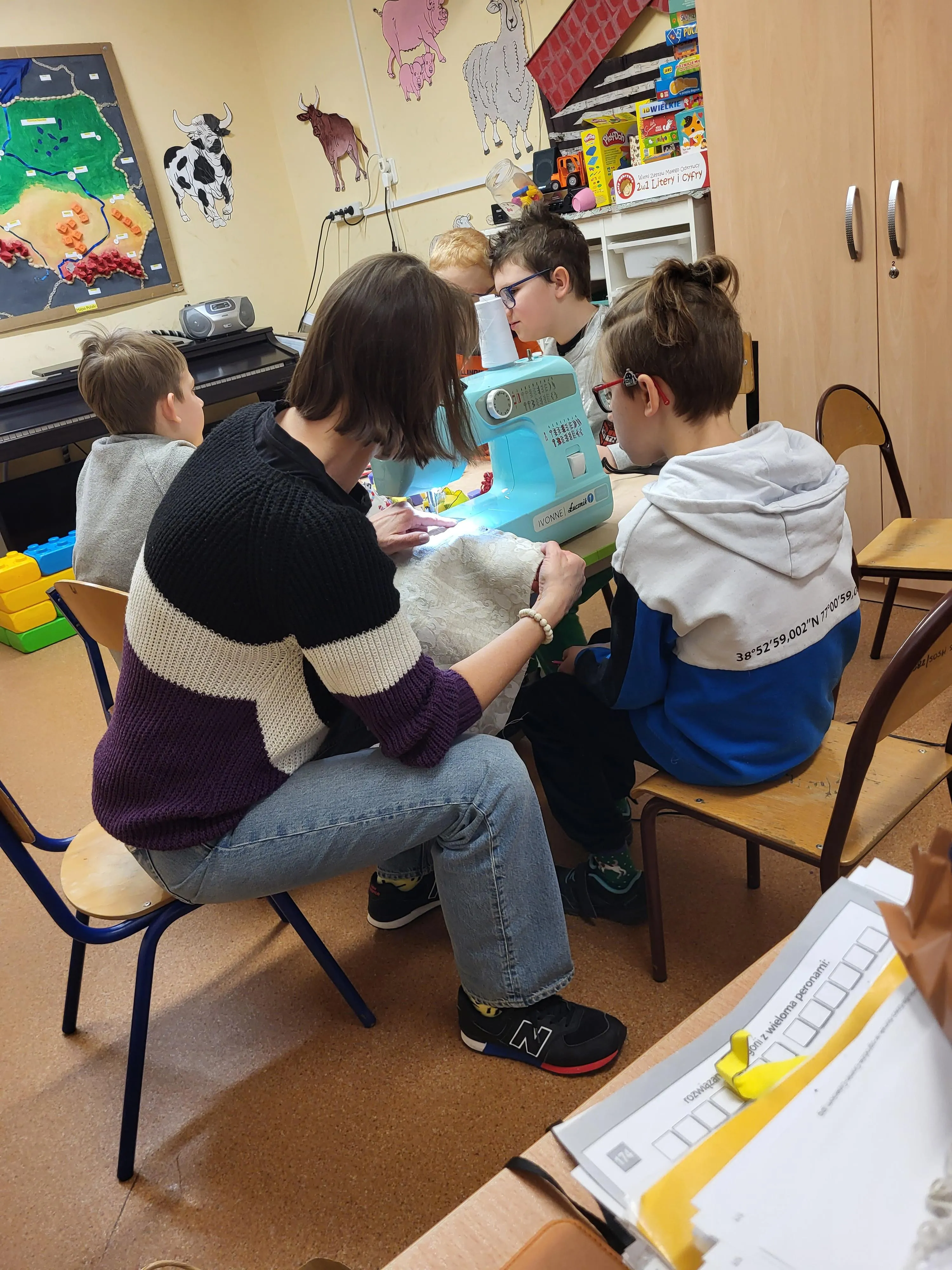 Na zdjęciu widać nauczycielkę oraz kilkoro dzieci siedzących przy stole w sali szkolnej. Wspólnie pracują przy maszynie do szycia. Dzieci obserwują i pomagają podczas szycia materiałowej torny. W tle widać szafki, zabawki oraz dekoracje na ścianach. 