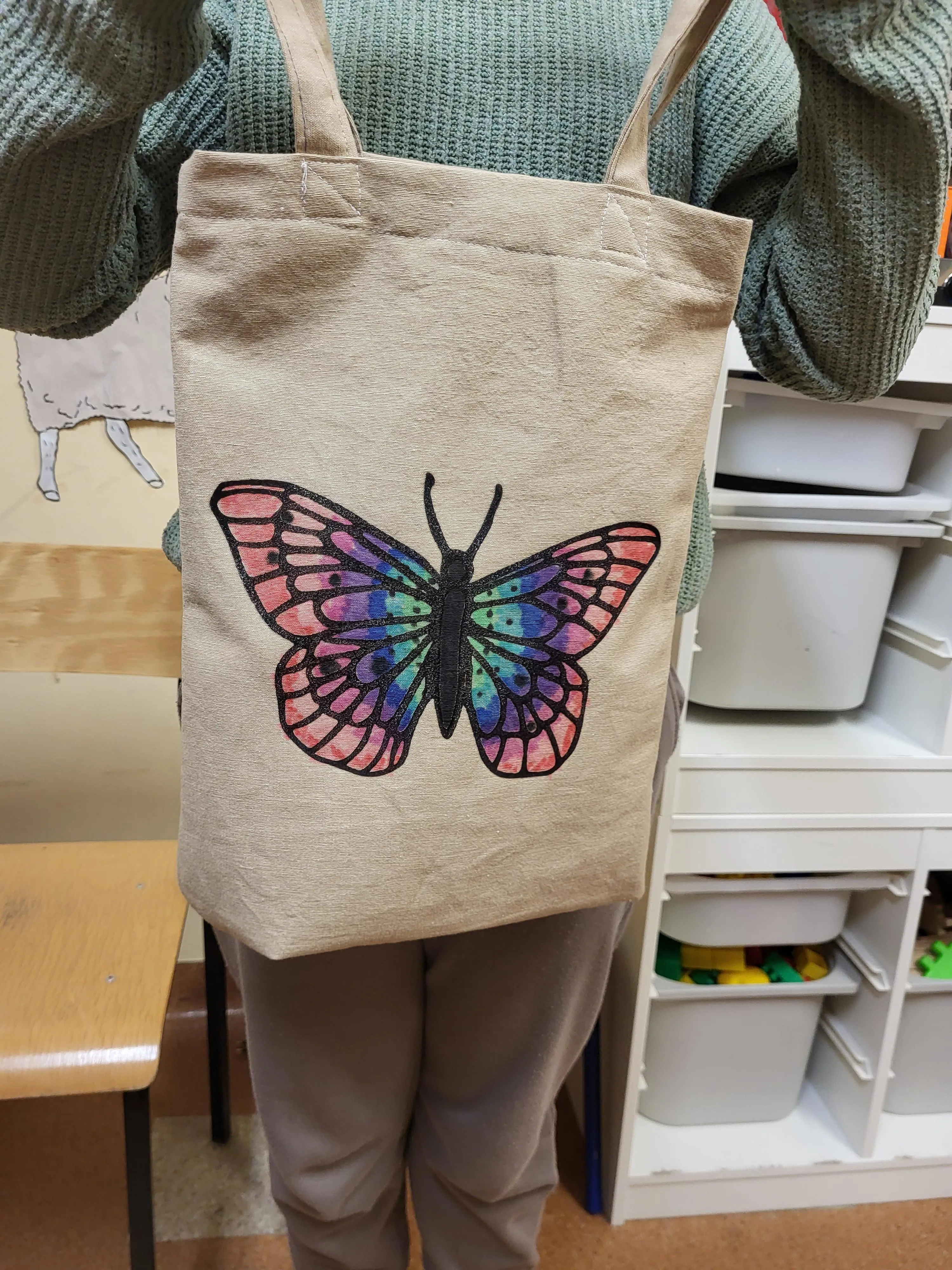 Na zdjęciu widać dziecko trzymajace materiałową torbę na ramię. Na środku torby znajduje się kolorowy rysunek pdzedstawiający motyla. Skrzydłą motyla zostały pokolorowane na różne kolory,iędzy innymi różowy, niebieski i fioletowy. 
