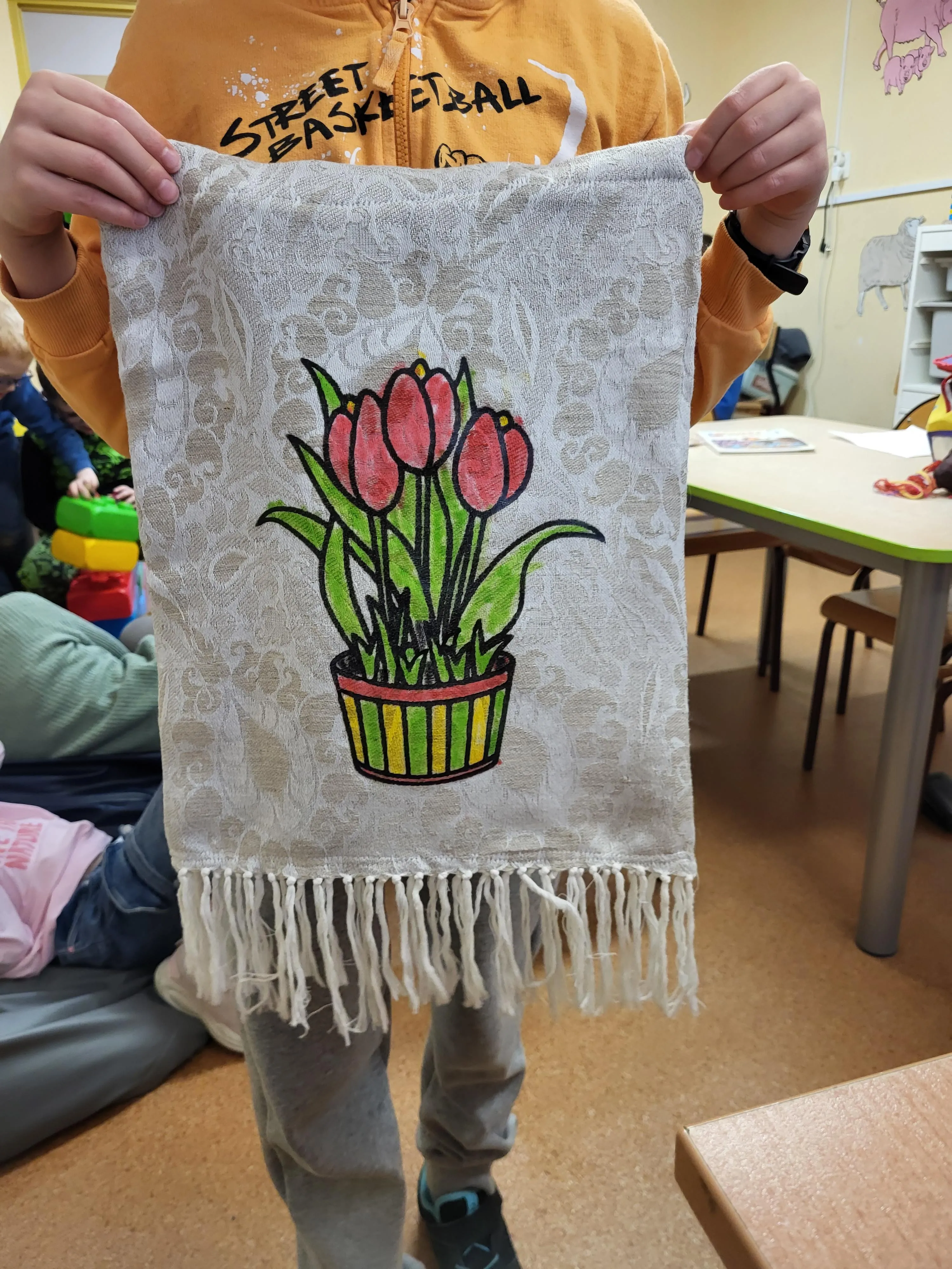 Na zdjęciu widać sylwetke dziecka trzymające uszyt a materiałową torbę. Na środku torby znajduje się kolorowy rysunek przedstawiający czerwone tulipany w doniczce. Nadruk został pokolorowany flamastrami. Torba ma jasny kolor oraz ozdobne frędzle na dole. 