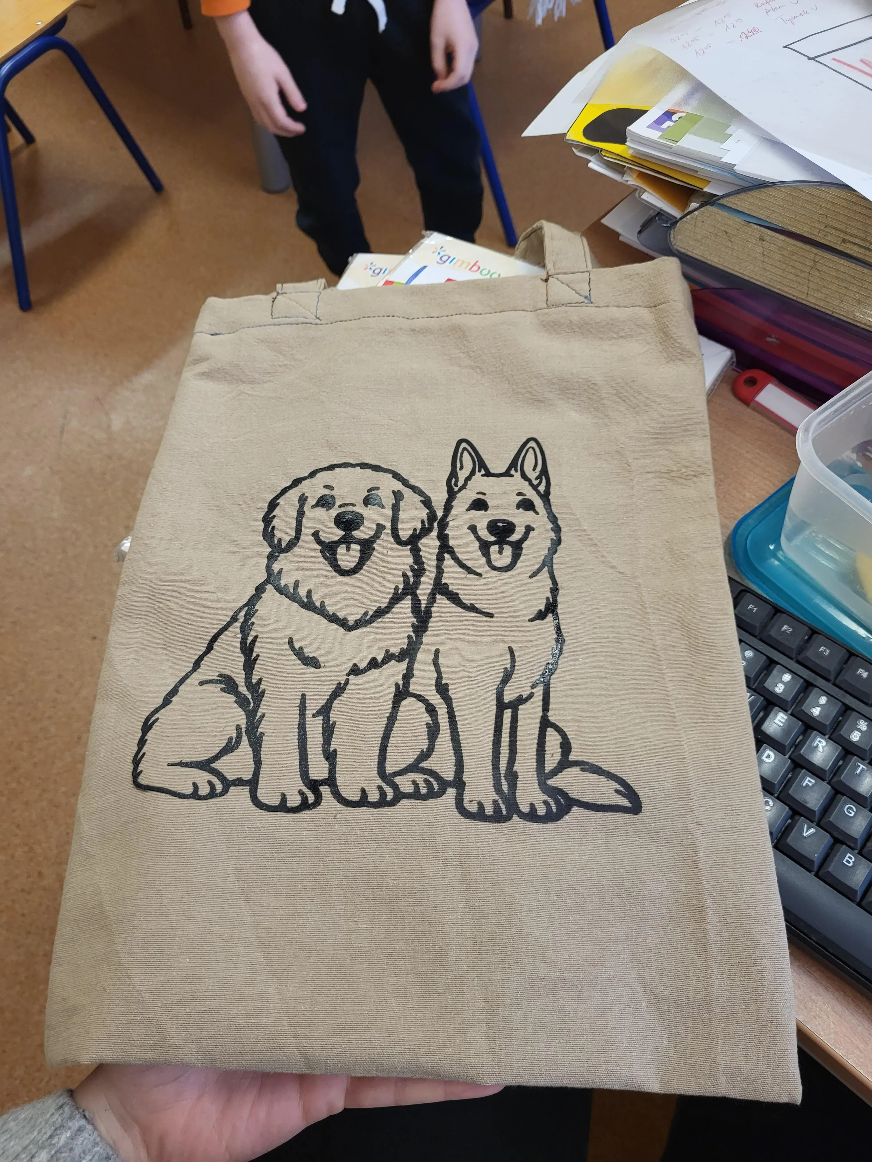 Na zdjęciu widać materiałową torbę. Na jej środku znajduje się czarny rysunek przedstawiający dwa siedzące psy. Torba ma jasny, naturalny kolor. Zdjęcie zostało wykonane w sali szkolnej. W tle widać biurko, klawiaturę komputera oraz przybory szkolne.