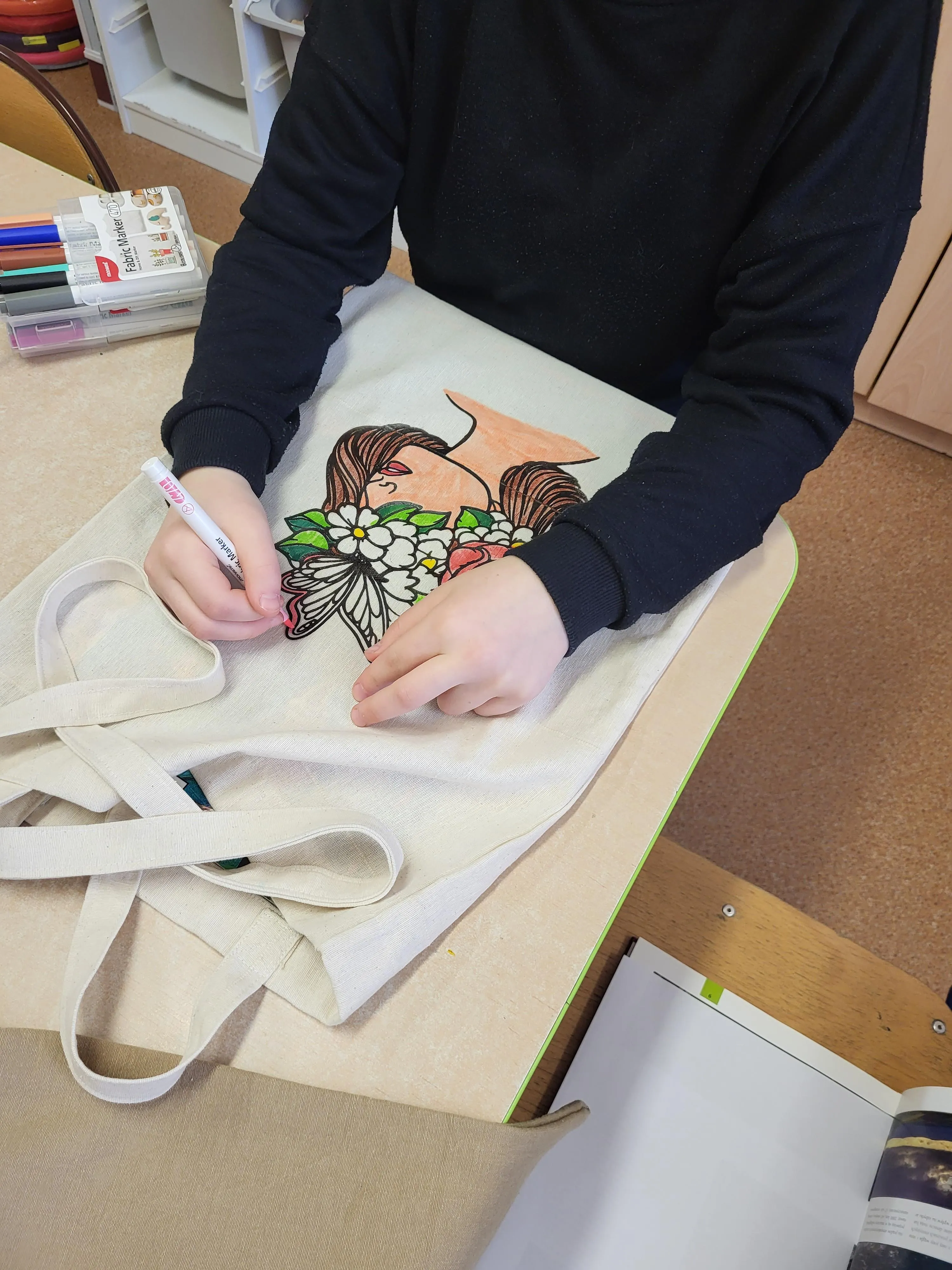 Na zdjęciu widać ucznia siedzącego przy stole i kolorującego nadruk na materiałowej torbie. Nadruk przedstawia postać z długimi włosami oraz kwiaty. Do kolorowania używane są kolorowe markery. 