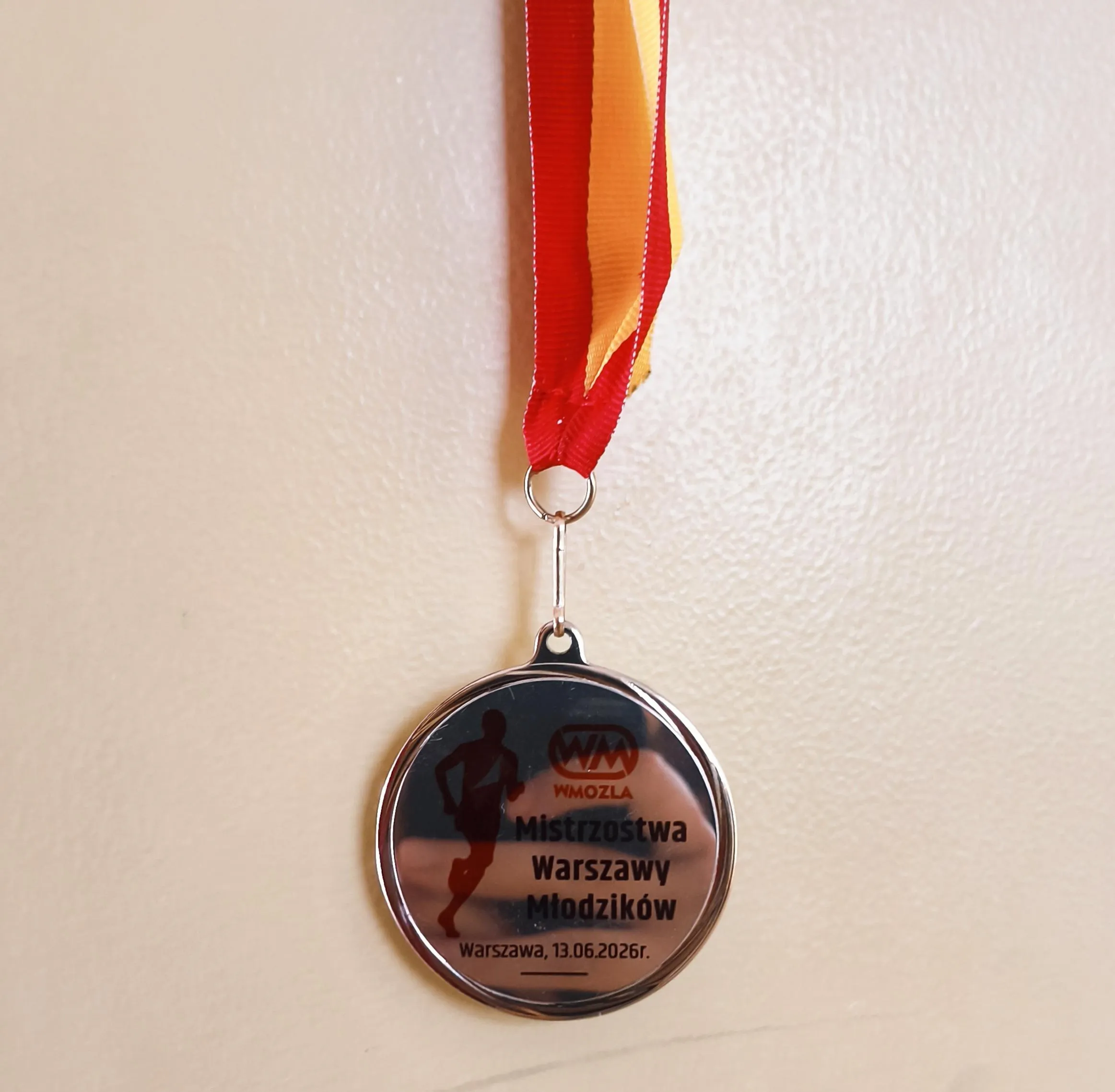 Medal zdobyty przez uczennicę podczas Mistrzostw Warszawy Młodzików, zawieszony na czerwono-żółtej wstążce.